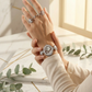 Hand wearing a silver watch with floral design on a marble surface with greenery