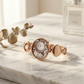 Rose gold bracelet with intricate design on a marble surface next to a bottle of perfume.