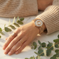 Hand wearing a gold cameo watch and ring with green leaves in the background