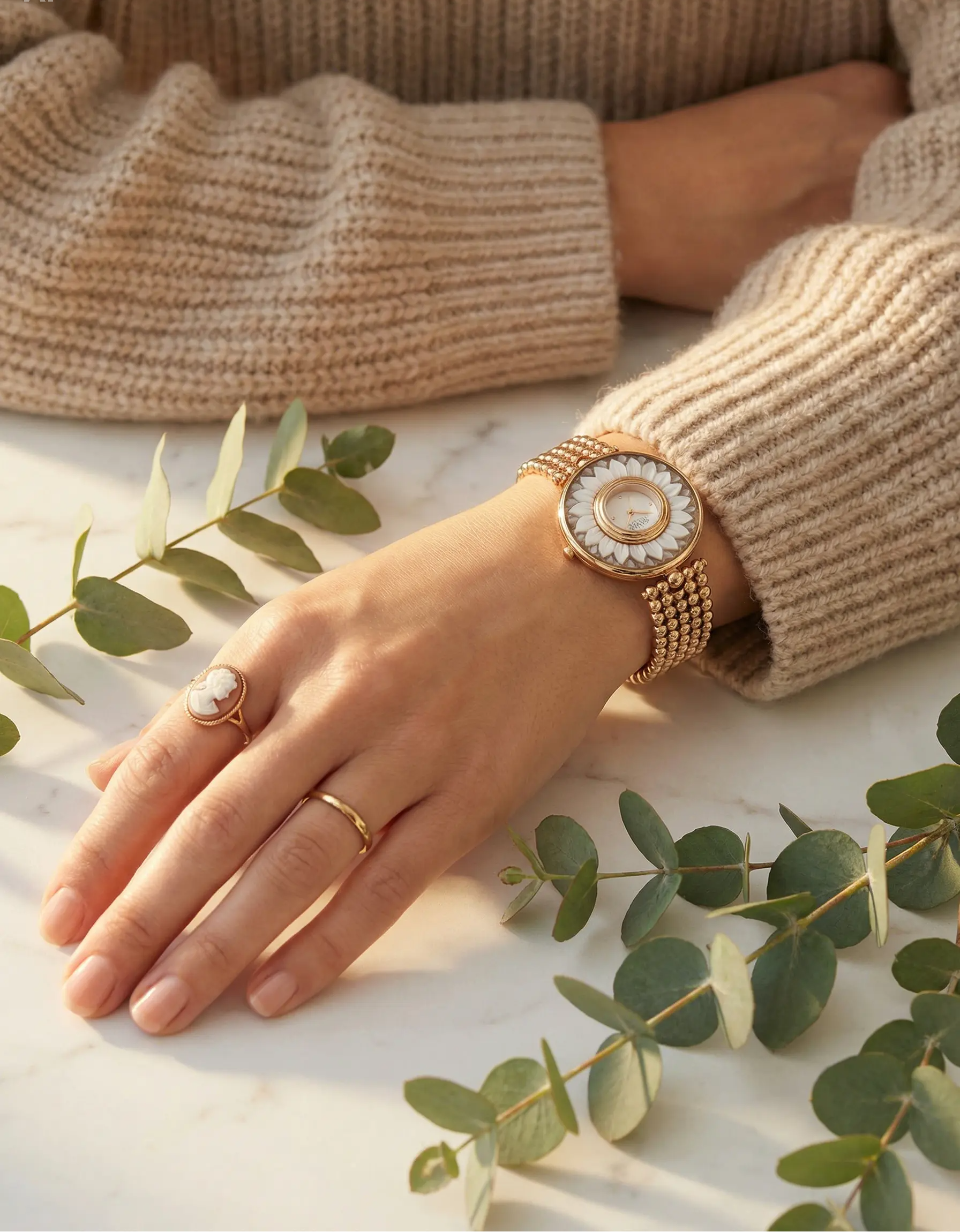 Hand wearing a gold cameo watch and ring with green leaves in the background