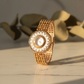 Gold cameo watch with intricate design on a reflective surface with leaves in the background