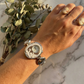 Hand wearing a decorative watch with a marble background and greenery