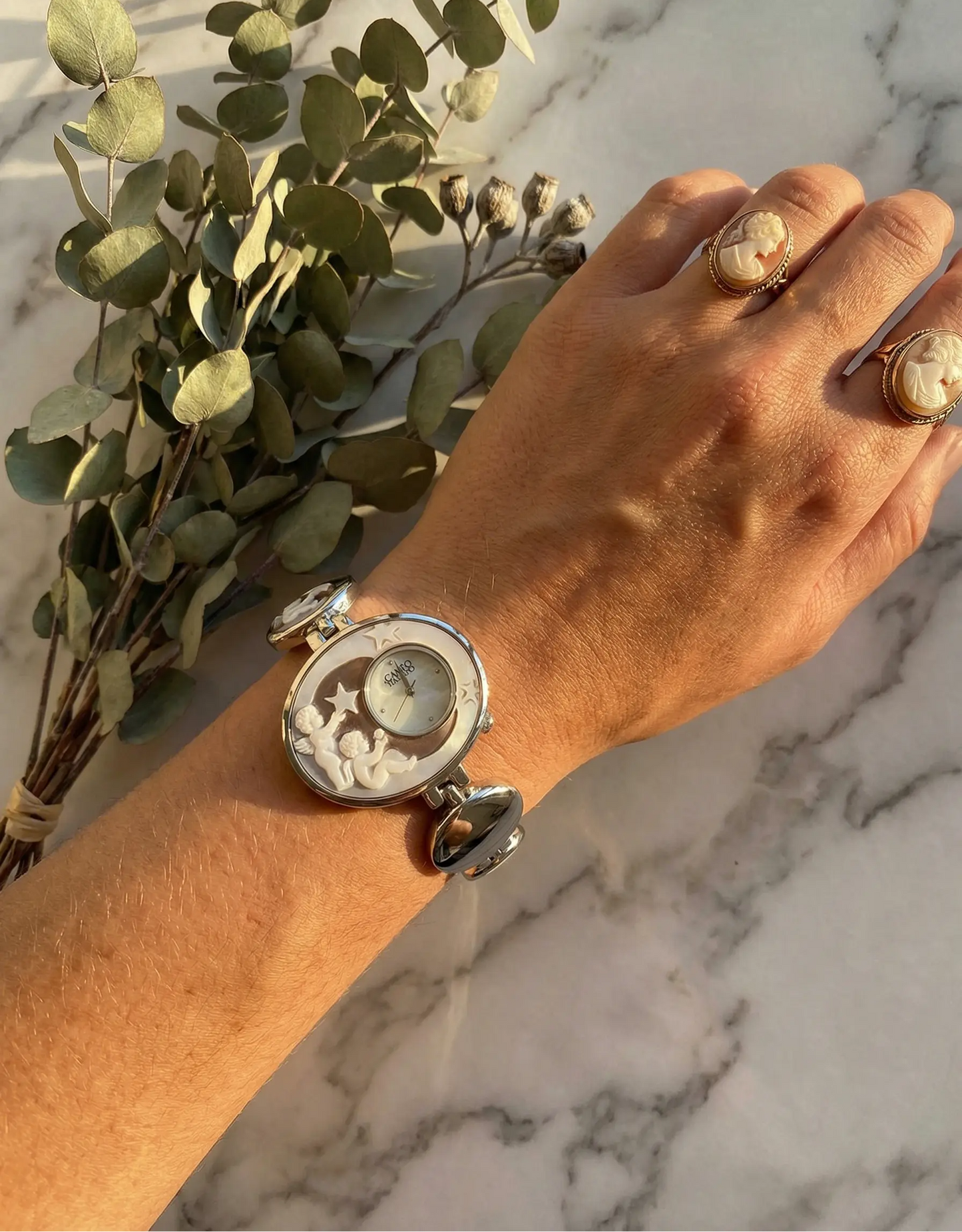 Hand wearing a decorative watch with a marble background and greenery