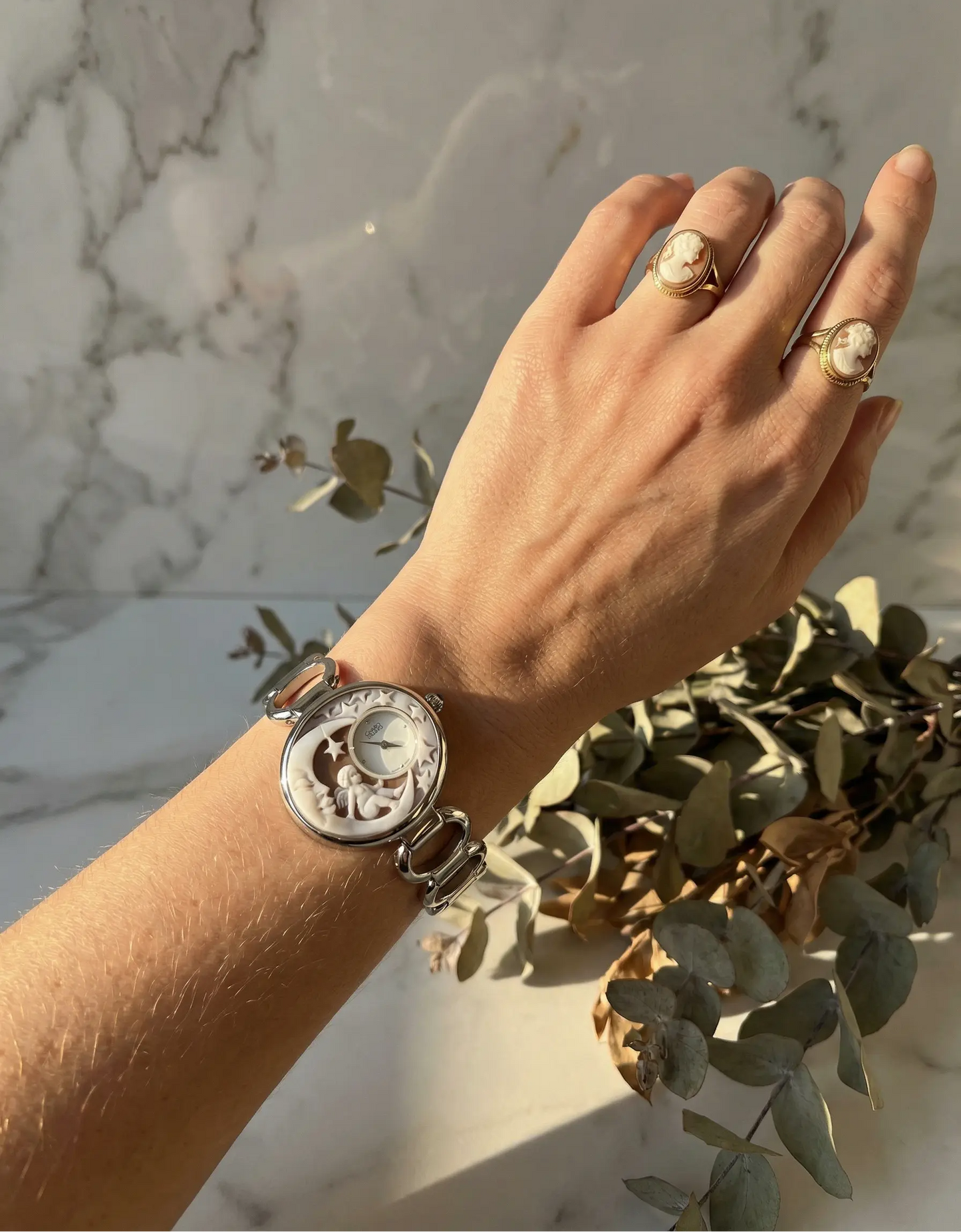 Hand wearing a silver watch with visible brand logo, against a marble wall background.