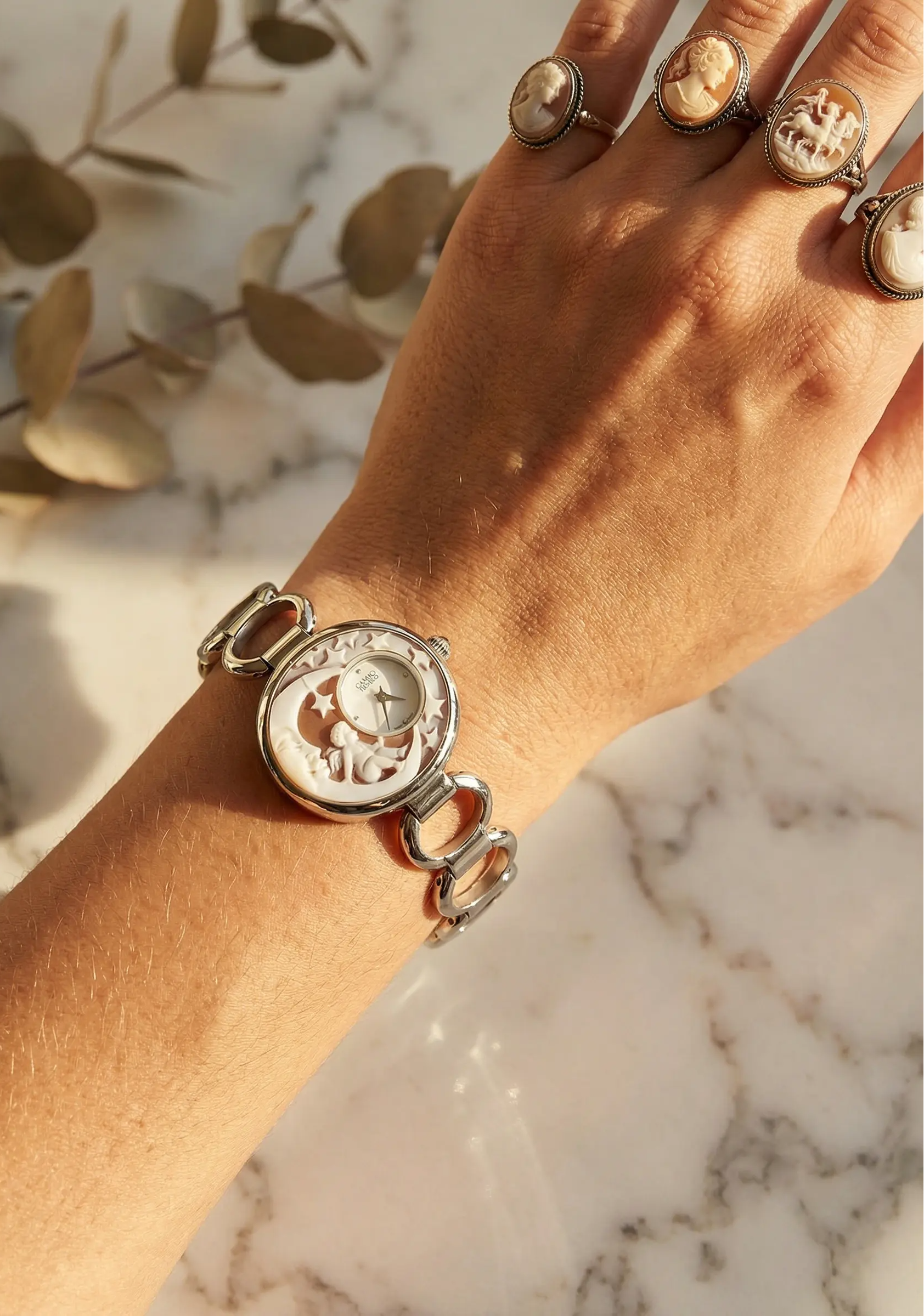 Hand wearing a silver bracelet with a circular design on a marble surface with eucalyptus leaves in the background.