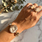 Hand wearing a gold bracelet and ring on a marble surface with greenery.
