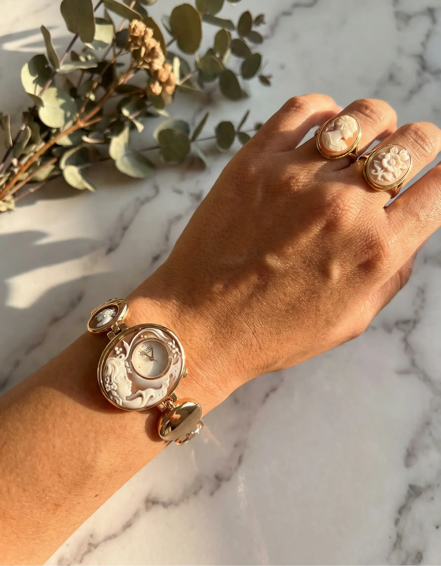 Hand wearing a gold bracelet and ring on a marble surface with greenery.