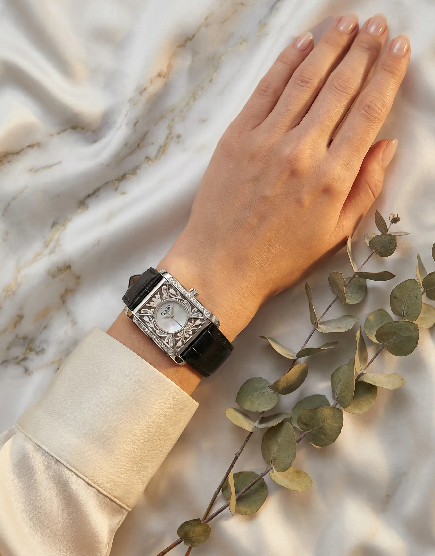Hand wearing an antique watch on a marble surface with green leaves.