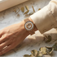 Hand wearing a gold watch with floral design on a marble surface with green leaves