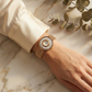 Hand wearing a gold watch with a decorative face on a marble surface with green leaves.