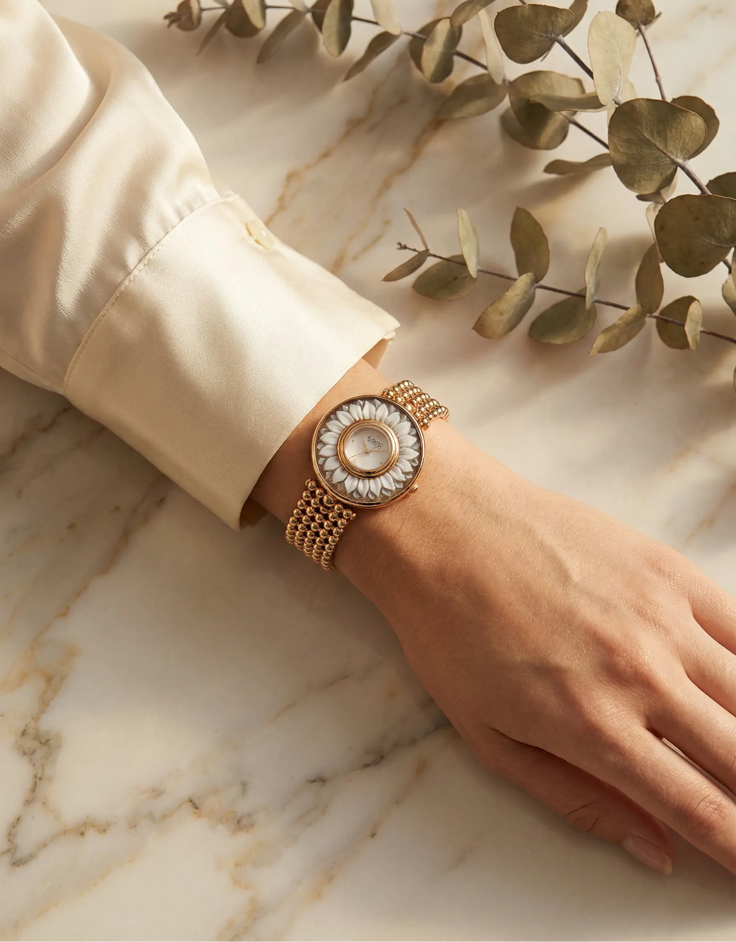 Hand wearing a gold watch with a decorative face on a marble surface with green leaves.