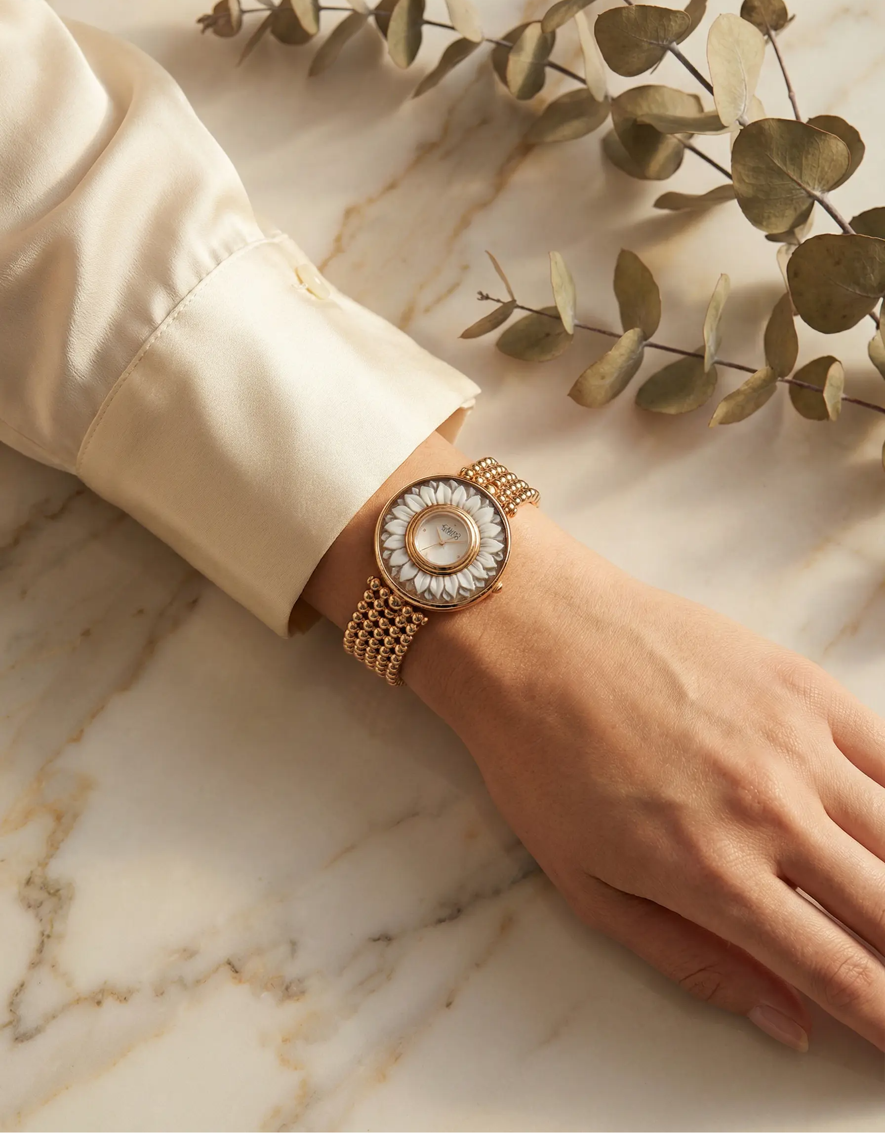Hand wearing a gold watch with a decorative face on a marble surface with green leaves.
