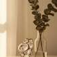 Silver bracelet with a decorative element next to a clear vase with eucalyptus leaves on a neutral background