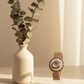 White vase with greenery and a decorative watch on a light surface.